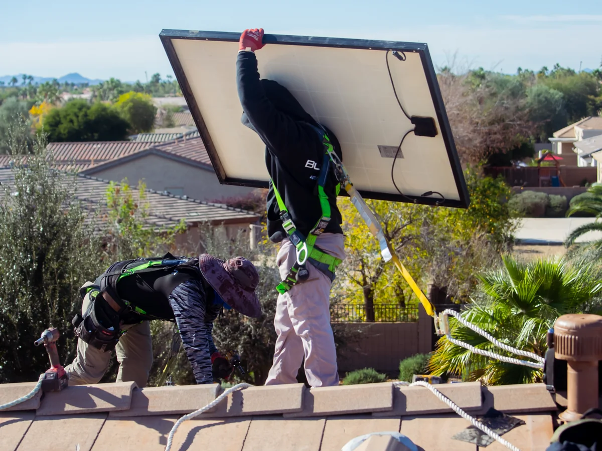 Two Blackout Electric technicians on a tile roof, one lifting a solar panel clear while the other works at the mounts