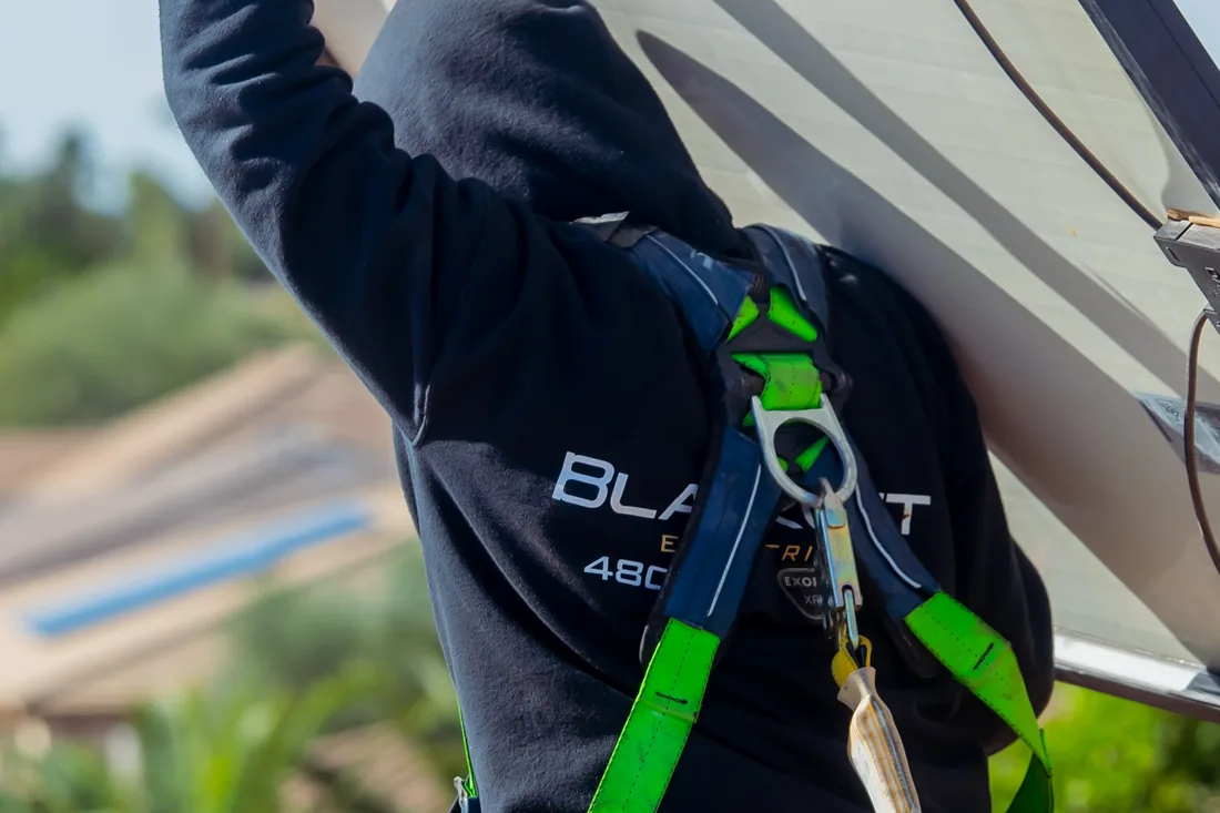 A harnessed Blackout Electric technician lifting a solar panel clear of a tile roof